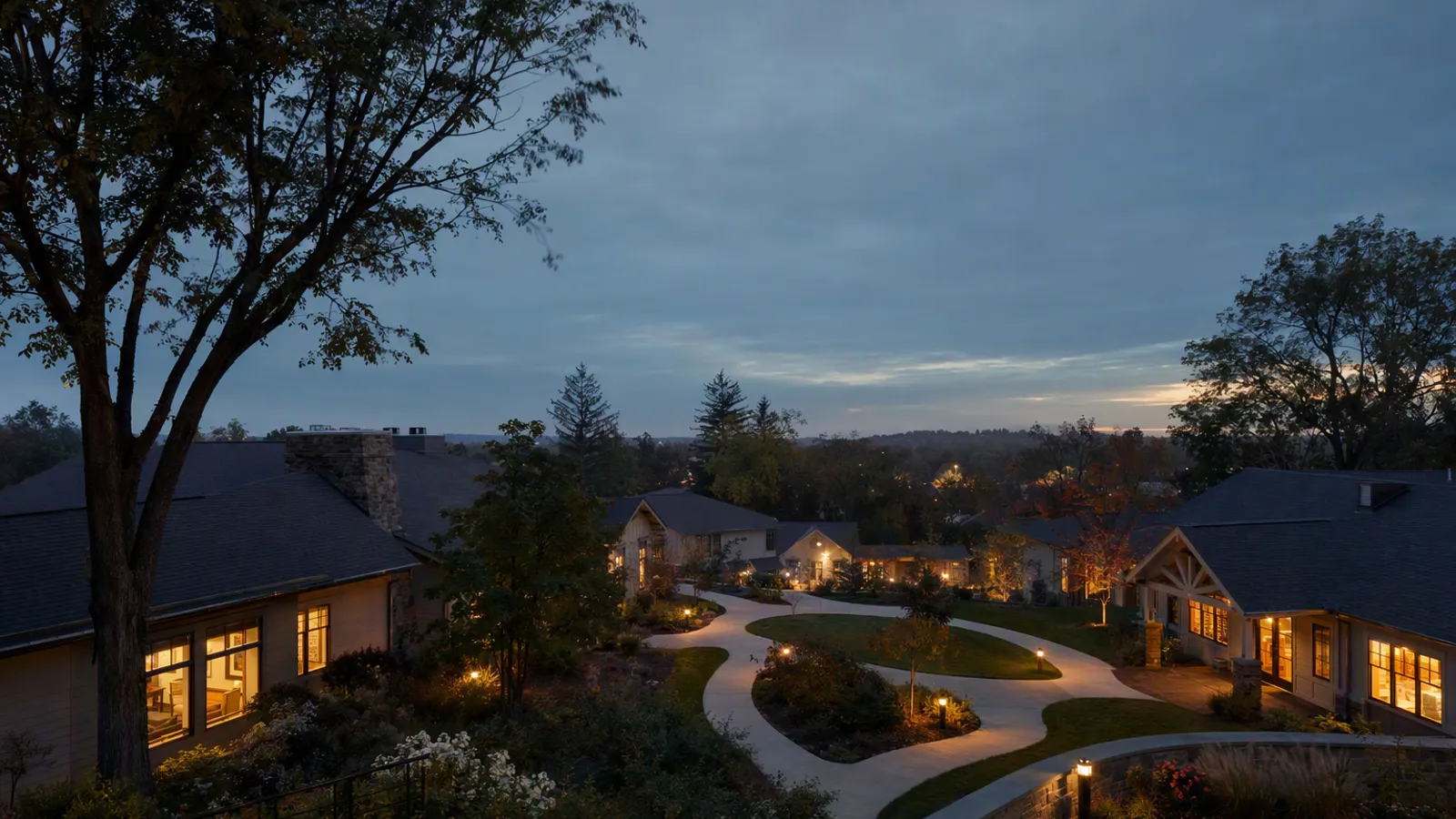 A senior-living community at dusk, warm lit windows around a landscaped courtyard and a lit garden path