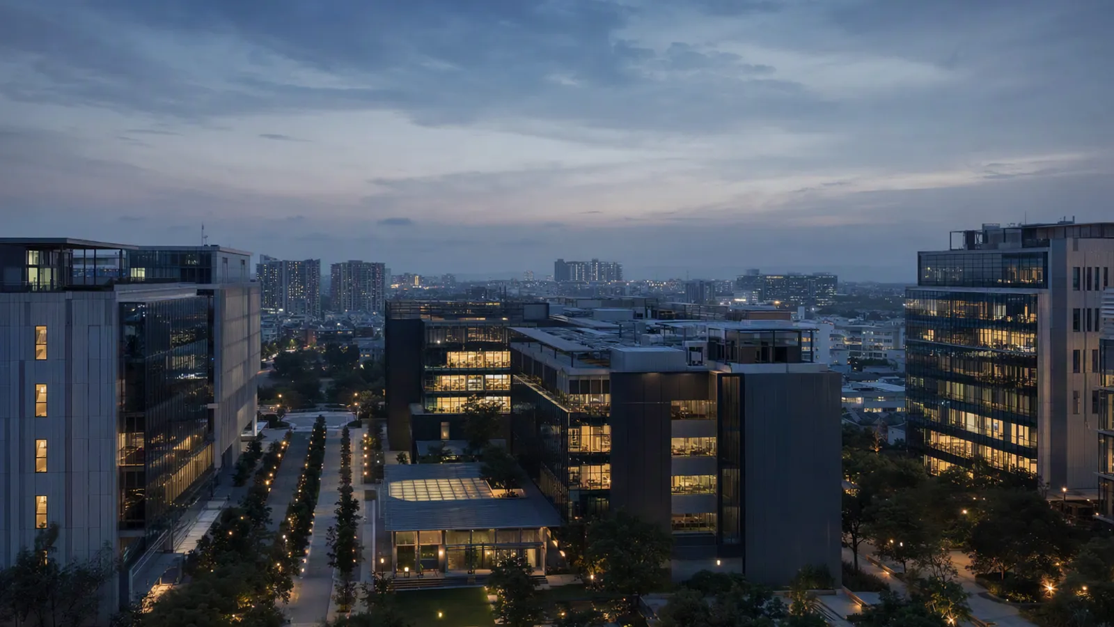 A commercial office campus at blue hour from above, glass towers with lit floors around landscaped courtyards
